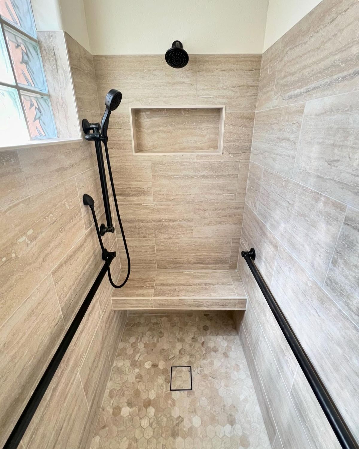 Walk-in shower with travertine tile, hex floor, niche, and black fixtures